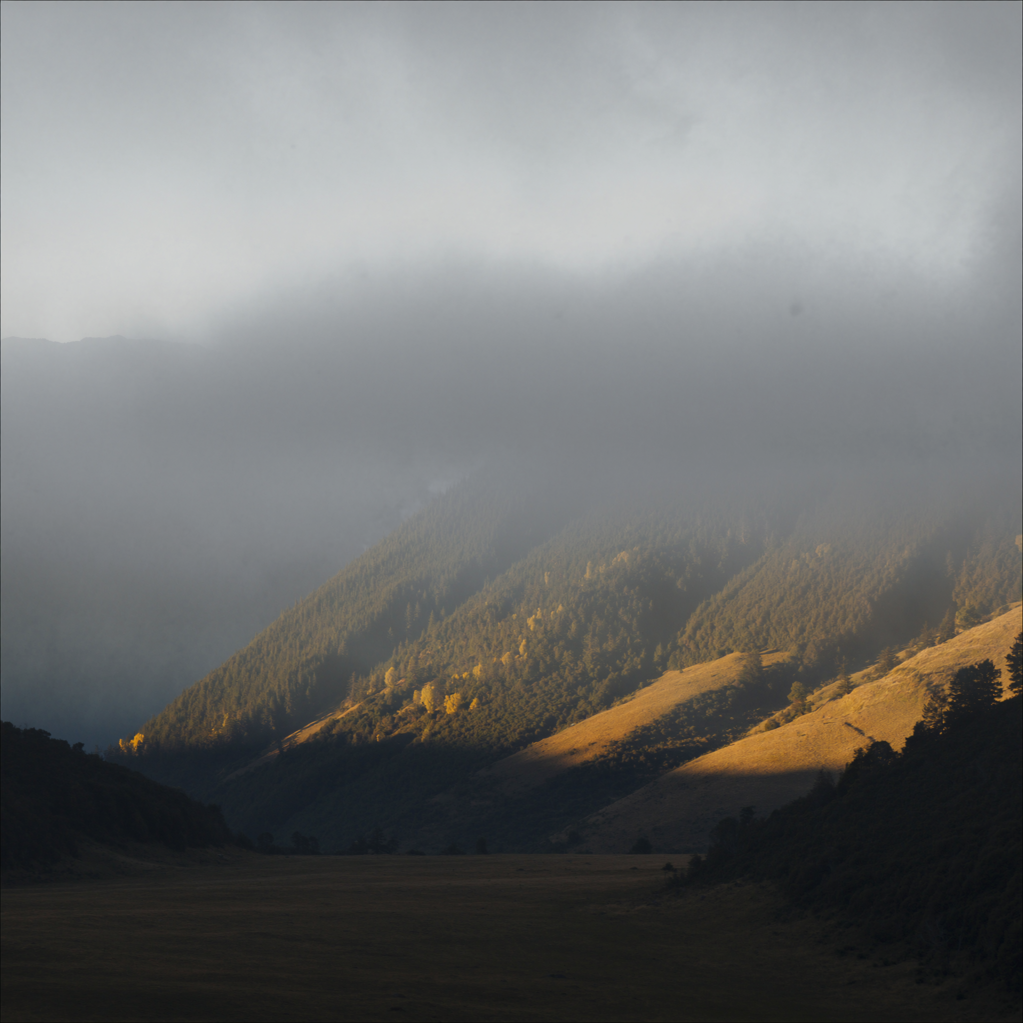 Alpines Bergtal am fruehen Morgen: Nebel weicht aus dem Talboden, die untere Hangflanke liegt in warmem Licht, der Gipfel steckt noch in der Wolke.